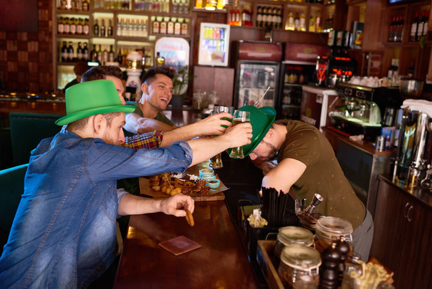 Группа веселых друзей, пьющих бокалы с пивом, сидя за барной стойкой и празднуя День Святого Патрика, бородатый бармен в зеленой шляпе котелка кладет голову под очки
 - Фото, изображение