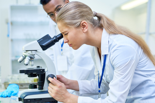 Side view portrait of young female scientist looking in microscope while working on medical research in science laboratory - Photo, Image