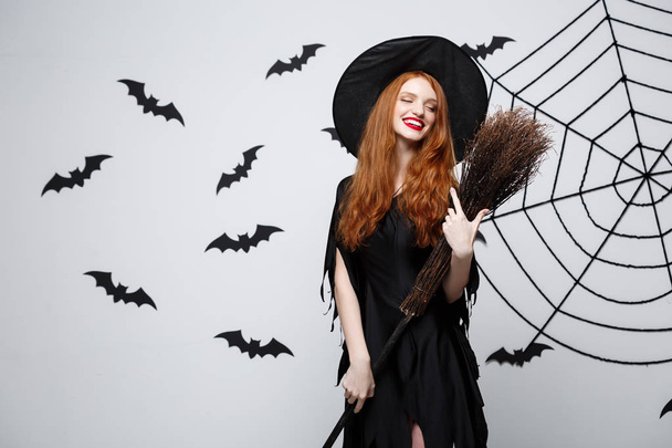 Halloween čarodějnice koncept - portrét krásné mladé čarodějnice s koště nad šedou zeď s bat a spider webové pozadí. - Fotografie, Obrázek