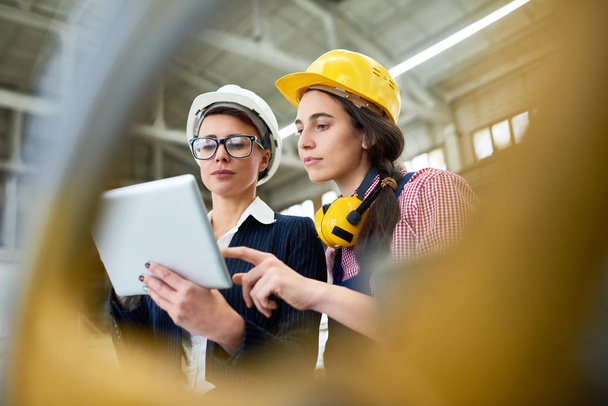 Retrato de dos ingenieras con sombreros de pie en un amplio taller de planta moderna usando tableta digital
 - Foto, Imagen