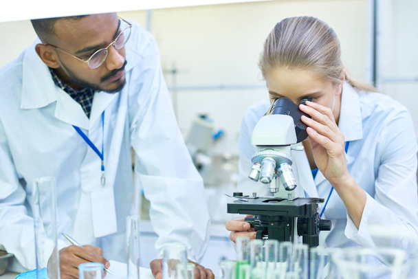 Porträt eines jungen Wissenschaftlers aus dem Nahen Osten, der Notizen auf Klemmbrett macht, während er im Labor an der medizinischen Forschung arbeitet, während seine Kollegin ins Mikroskop schaut - Foto, Bild