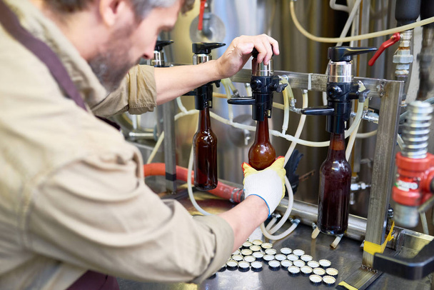 Eldiven giymiş sakallı makine operatörünün görünümünü profil ve dolum bira şişeleri modern bira fabrikası işyerinde sarılı iken tankından önlük - Fotoğraf, Görsel