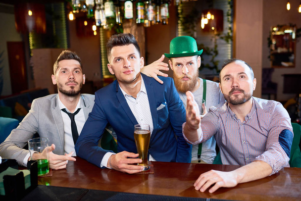 Spending St. Patrick's Day with friends: group of bearded men watching rugby match and enjoying beer while sitting at bar counter, one of them wearing leprechaun costume - Foto, Imagem