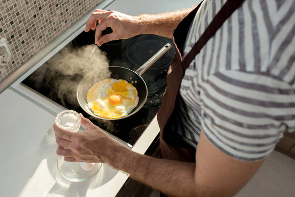 高角度が日光に照らされてモダンなキッチンで朝食を作っている間にフライパンで卵を塩漬け男性の手のクローズ アップ - 写真・画像