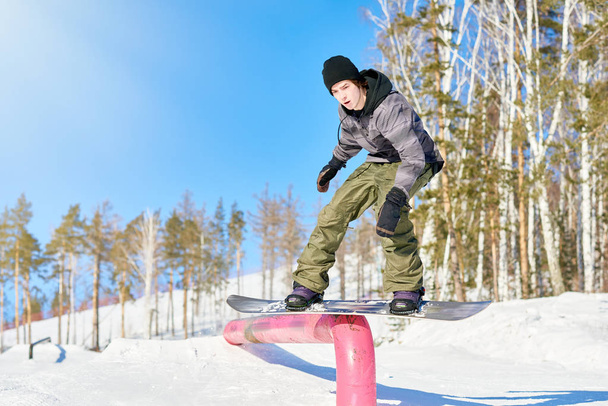 tiro de acción de cuerpo entero del joven realizando acrobacias de snowboard deslizándose por la barandilla metálica a la luz del sol en la estación de esquí
 - Foto, Imagen