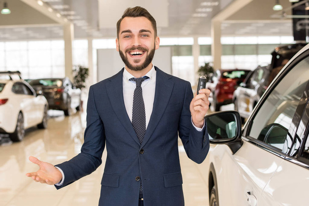 Porträt eines gutaussehenden Autoverkäufers, der Autoschlüssel in der Hand hält und fröhlich in die Kamera lächelt, die im Showroom eines Luxus-Autohauses steht - Foto, Bild