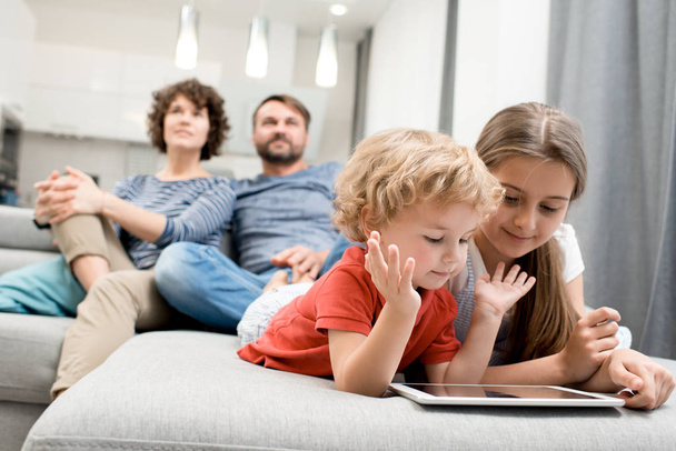 Mooi klein meisje en haar krullend broer liggend op gezellige Bank en speelspel op digitale tablet terwijl hun ouders verpakt in Tv kijken, interieur van studio appartement op de achtergrond - Foto, afbeelding