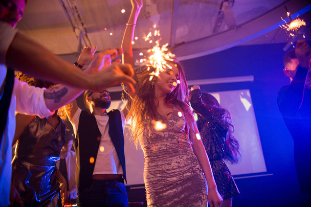 Group of trendy young people celebrating holiday in nightclub burning flaming sparklers in middle of dance floor, focus on dancing beautiful girl wearing  glittering velvet party dress - Photo, Image