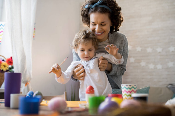 Retrato de madre joven feliz ayudando a pintar huevos de Pascua mientras se preparan para las vacaciones en casa
 - Foto, Imagen