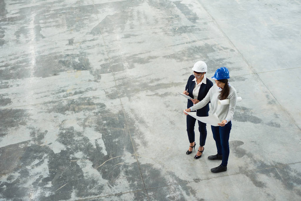 High angle view of two female engineers wearing hardhats examining floor plans in workshop of modern factory - Фото, изображение