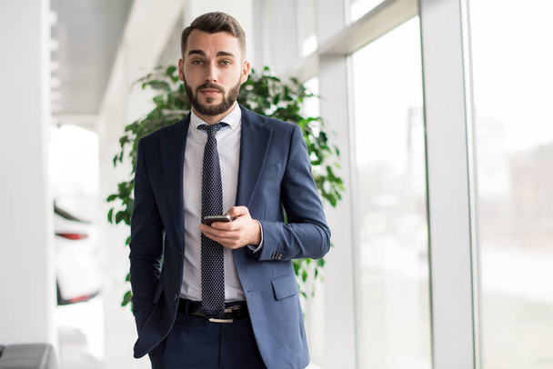 Portrait of handsome bearded businessman holding smartphone and looking at camera standing by window in modern office, copy space - Photo, Image