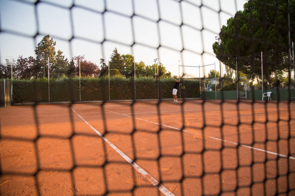 Professional tennis player man playing on the court in the afternoon seen through the net.  - Photo, Image