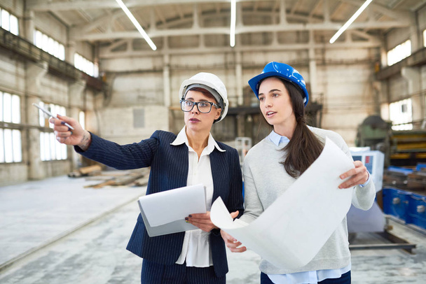 Retrato de dos ingenieras vestidas con sombreros de pie en un amplio taller de planta moderna señalando y sosteniendo el plan de construcción
 - Foto, imagen