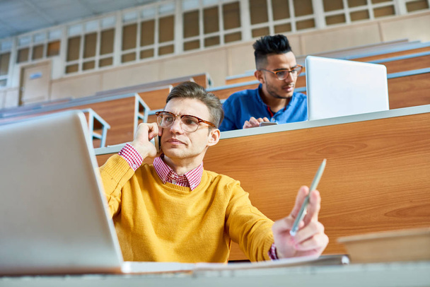 Niski kąt widzenia dwóch studentów, jeden z nich Środkowo Wschodniej, siedząc na biurka w nowoczesne audytorium College i przygotowanie do klasy, skupić się na młodego człowieka za pomocą laptopa i mówienie przez smartfona - Zdjęcie, obraz