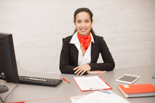 Portrait of cheerful mixed-race woman wearing black and red uniform sitting at desk in modern office smiling happily at camera, copy space - Photo, Image