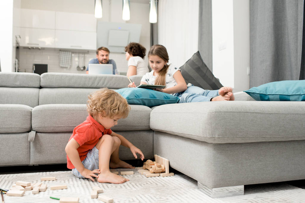 entzückender kleiner Junge sitzt auf Teppich und spielt mit Holzspielzeug, während seine ältere Schwester auf dem Sofa liegt und skizziert, ihre Eltern unterhalten sich angeregt in der Küche - Foto, Bild
