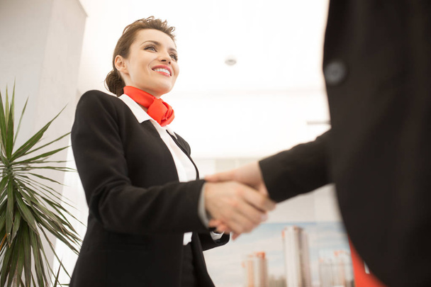 Низький кут зору на гарненьку бізнес-леді тремтить руками з клієнтом, посміхаючись щасливо стоячи в сучасному офісі
 - Фото, зображення