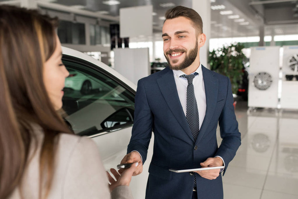 Ritratto di affascinante venditore di auto dando chiavi a giovane donna in piedi accanto a bianco lucido auto di lusso in concessionaria showroom
 - Foto, immagini