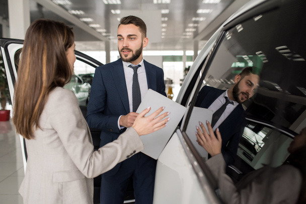 Portrait of handsome car salesman showing white luxury car to young woman in dealership showroom - Photo, Image
