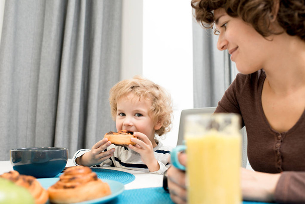 Enjoying breakfast in bosom of family: adorable curly boy looking at camera while eating appetizing bun, his pretty middle-aged mother with warm smile keeping eye on him - Фото, изображение