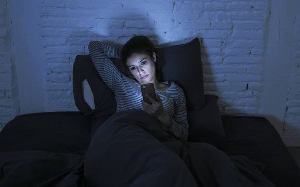 portrait of young beautiful latin woman using mobile phone late night sleepless lying in bed in the dark in smartphone and internet dating app addiction  - 写真・画像