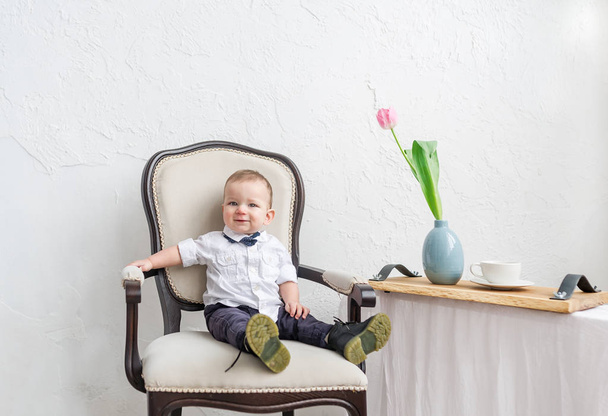  Schattige babyjongen onder 1 jaar oude dragen van stijlvolle kleding in vintage stoel in huis kamer zit. - Foto, afbeelding