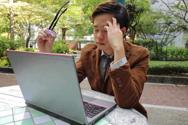 Joven hombre de negocios cansado sosteniendo gafas y sufrimiento ocular en frente de la computadora portátil de pantalla en el parque al aire libre
. - Foto, imagen