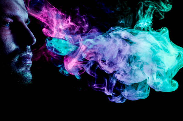 A young man exhales a cloud of colored smoke of blue and pink color on a black isolated. Harm to smoking on the example of a young mugger - Fotoğraf, Görsel