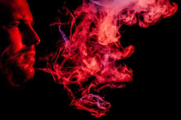 A young man exhales a cloud of colored smoke of  red color on a black isolated. Harm to smoking on the example of a young mugger - Foto, Imagen