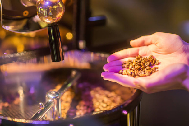 Foto recortada de manos de un hombre sosteniendo granos de café aromáticos recién tostados sobre una máquina moderna utilizada para asar granos
. - Foto, Imagen