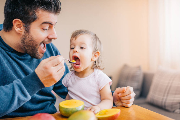 close-up of young happy father feeding cute little girl with fresh mango at home  - Foto, Bild