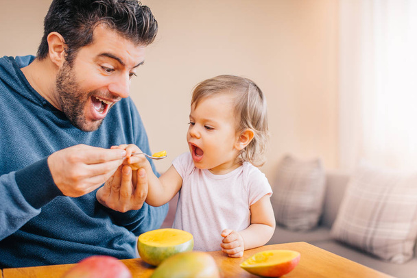 close-up of young happy father feeding cute little girl with fresh mango at home  - Foto, afbeelding