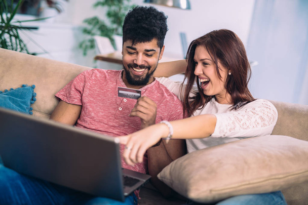 Homem usando laptop e mulher segurando cartão de crédito. Casal jovem compras on-line com cartão de crédito em casa
 - Foto, Imagem