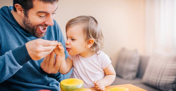 close-up of young happy father holding spoon and feeding cute little girl with fresh mango at home  - Foto, imagen