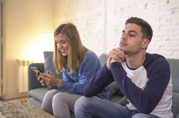 young couple at home sofa couch with woman internet and mobile phone addiction ignoring her boyfriend feeling sad jealous frustrated upset - Foto, imagen