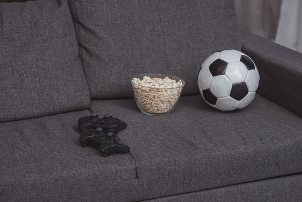 bowl with popcorn, football ball and gamepads on sofa - Fotó, kép