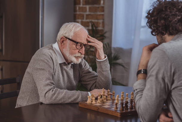 side view of adult son and senior father thinking while playing chess at home - Foto, Imagem