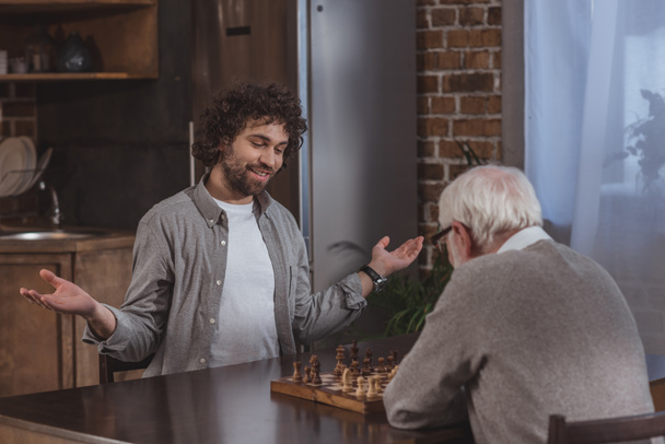 adult son gesturing while playing chess with senior father at home - Foto, afbeelding