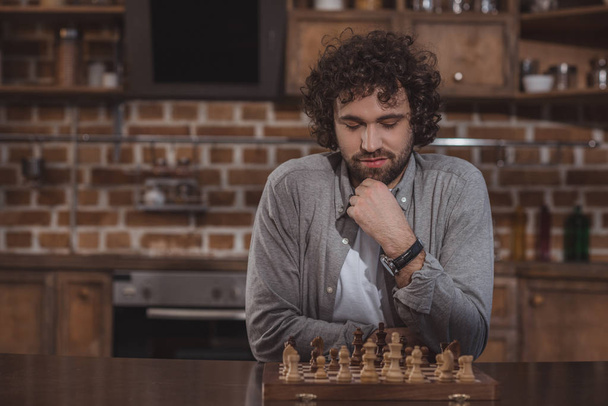 hombre guapo pensando y mirando tablero de ajedrez en casa
 - Foto, Imagen