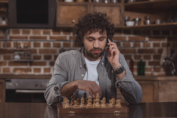 przystojny mężczyzna rozmawia przez smartfona i gra w szachy w domu - Zdjęcie, obraz