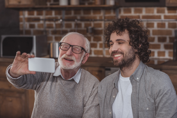 sorridente figlio adulto e padre anziano prendendo selfie in cucina
 - Foto, immagini