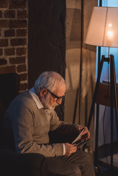 Seitenansicht eines gutaussehenden Mannes mit grauen Haaren, der im Sessel sitzt und zu Hause alte Fotos betrachtet - Foto, Bild
