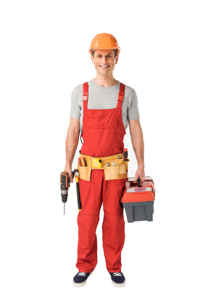 Cheerful construction worker in uniform holding toolbox and drill isolated on white - Photo, Image