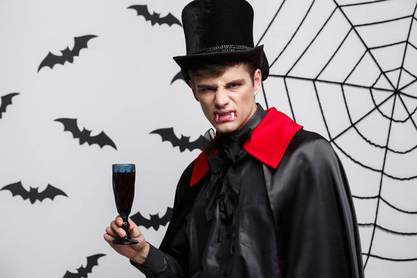 Vampir-Halloween-Konzept - Porträt eines hübschen kaukasischen Vampirs, der gerne blutigen Rotwein trinkt. - Foto, Bild
