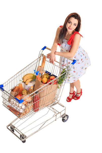 High angle view of girl smiling at camera while pushing a shopping cart full with groceries isolated on white background. - Foto, afbeelding