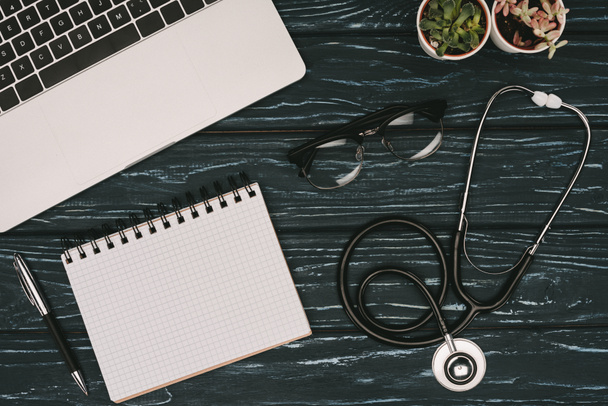 bovenaanzicht van gearrangeerde laptop, lege notebook, brillen en stethoscoop op donkere houten tafelblad - Foto, afbeelding