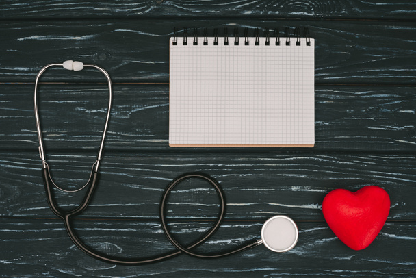 vlakke leggen met gearrangeerde rood hart, stethoscoop en lege notebook op donkere houten tafelblad, wereld gezondheid dag concept - Foto, afbeelding