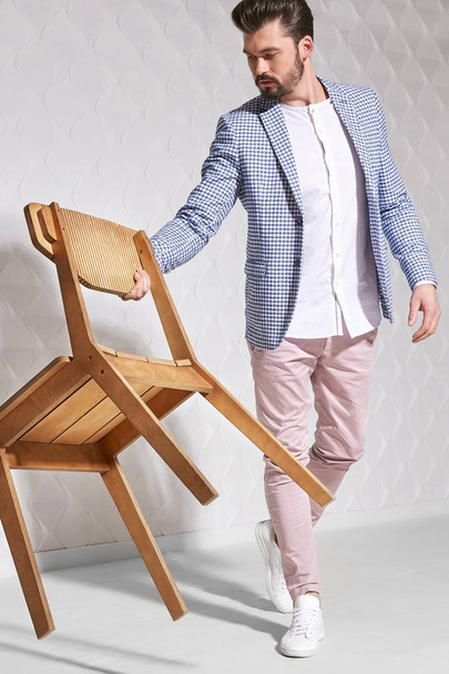 Fashion portrait of handsome male model with dark hair and beard wearing light pants, white shirt, plaid jacket and white shoes with chair near him - Fotó, kép