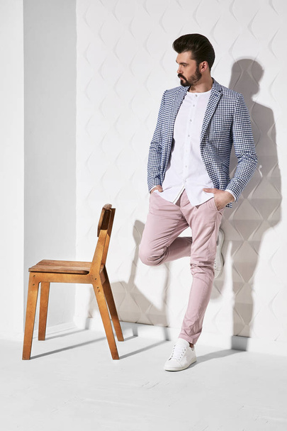 Fashion portrait of handsome male model with dark hair and beard wearing light pants, white shirt, plaid jacket and white shoes with chair near him - Fotó, kép