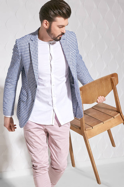Fashion portrait of handsome male model with dark hair and beard wearing light pants, white shirt, plaid jacket with chair near him - Foto, Imagen